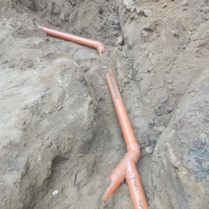 Pomarańczowe rury kanalizacyjne ułożone w wykopie ziemnym, widoczne złącza i fragmenty rur. Wykop w ziemi gliniastej, surowy stan instalacji.