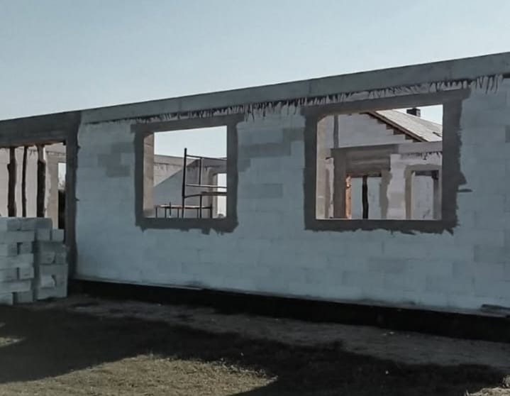 Ściana budynku w trakcie budowy z widocznymi otworami okiennymi i stertą bloczków. Widoczne rusztowania wewnątrz budynku. Surowy stan, wczesny etap prac.