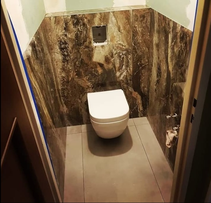 Wykończona łazienka z podwieszaną toaletą, brązowe płytki imitujące marmur na ścianach, szare płytki na podłodze. Widok zza futryny drzwi.