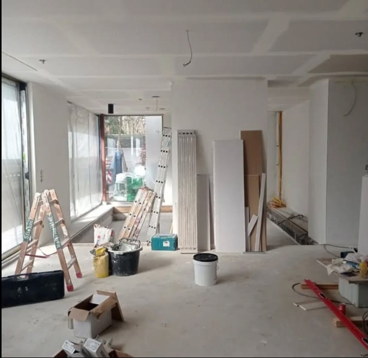 Wnętrze w trakcie remontu: widoczne drabiny, narzędzia, płyty gipsowe i szpachlowane sufity. Duże okno z widokiem na zewnątrz. Etap prac wykończeniowych.