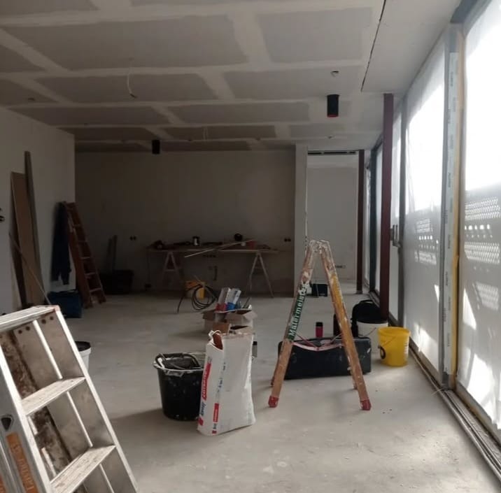 Wnętrze w trakcie prac wykończeniowych: sufit podwieszany z płyt gipsowych, drabiny, narzędzia i materiały budowlane. Widok na przestrzeń z oknami.