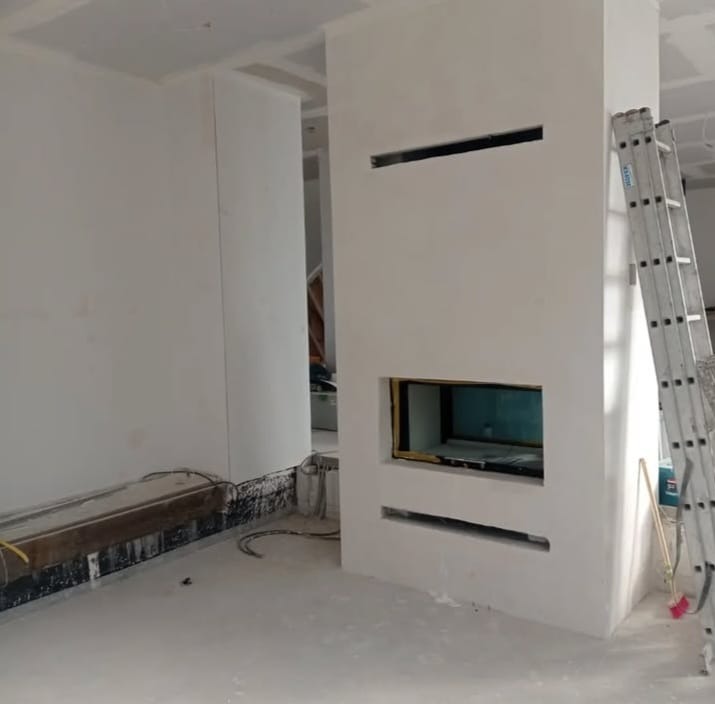 Wnętrze w trakcie wykańczania: białe ściany, wnęka kominkowa z widoczną instalacją, drabina oparta o ścianę. Widoczne przewody i elementy konstrukcyjne.