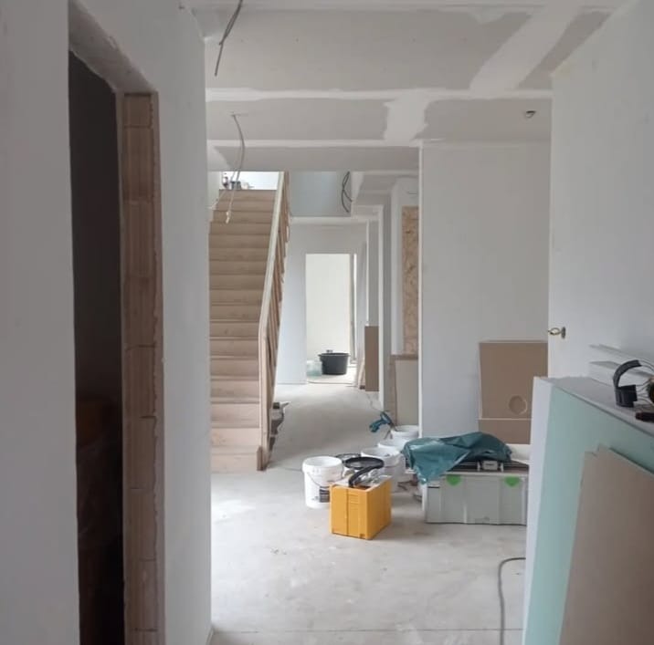 Wnętrze w trakcie prac wykończeniowych: surowe ściany, schody drewniane, widoczne narzędzia i materiały budowlane. Perspektywa z korytarza.