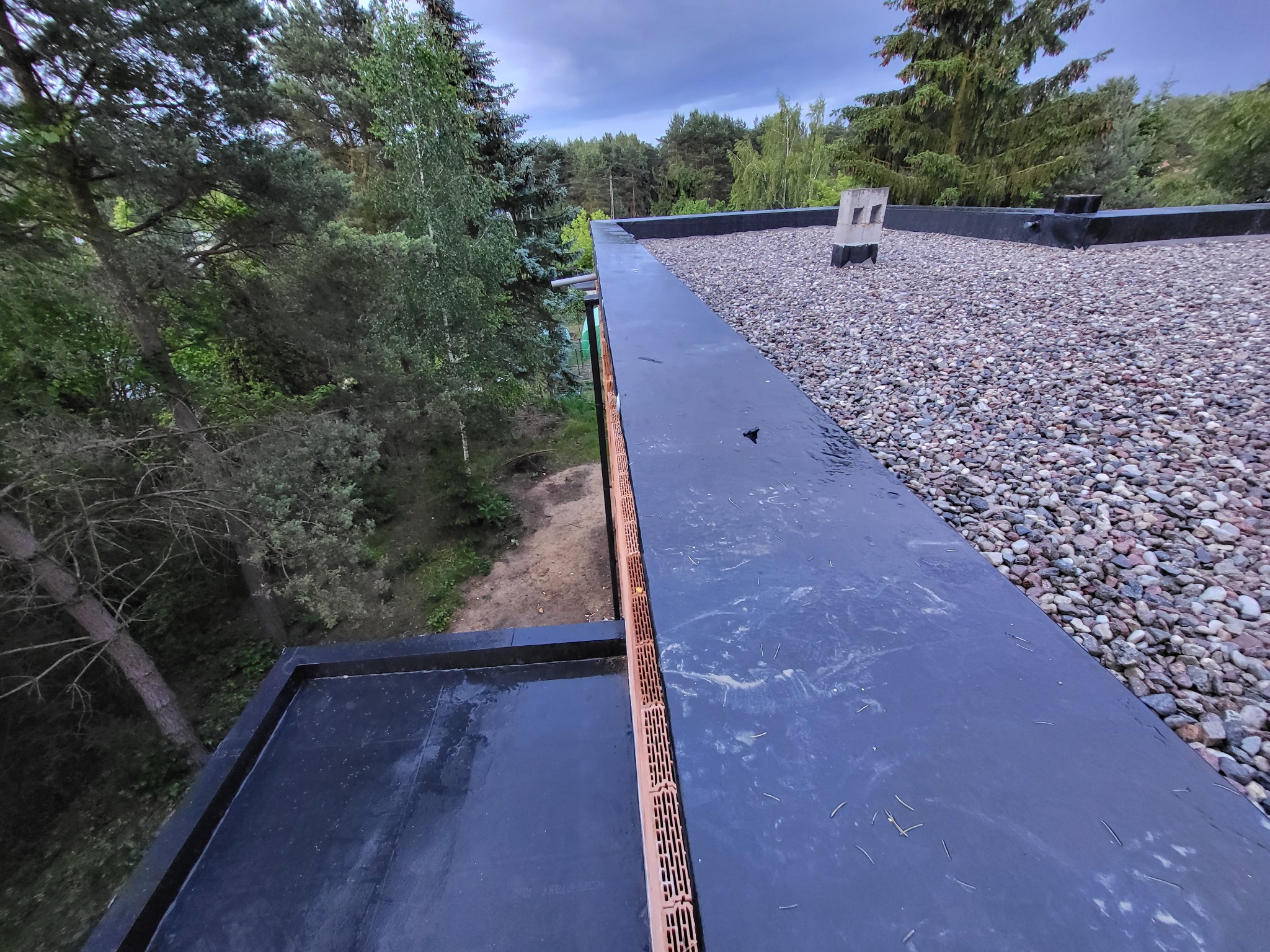 Wykończony płaski dach z czarną membraną i żwirem, widok z góry na tle zieleni. Widoczna ceramiczna kratka wentylacyjna. Dach po deszczu, mokra powierzchnia.