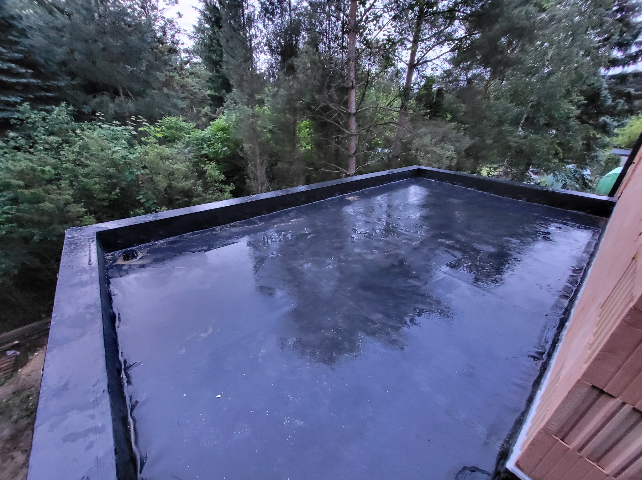 Wykończony, płaski dach pokryty czarną membraną bitumiczną, z widocznymi detalami obróbki krawędzi i fragmentem muru z cegły ceramicznej w tle zieleni.