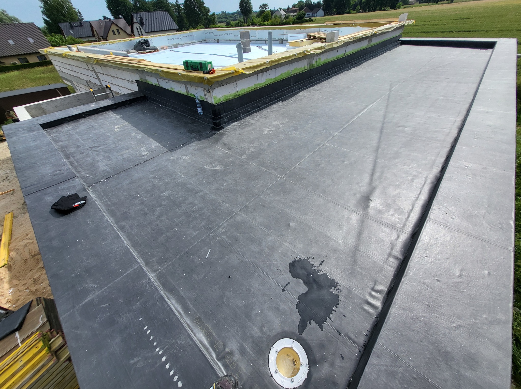 Czarna, płaska powierzchnia dachu z widocznymi warstwami izolacji i elementami konstrukcyjnymi. Na dachu widoczne narzędzia i ślady po pracy, w tle zielone pola i zabudowa.