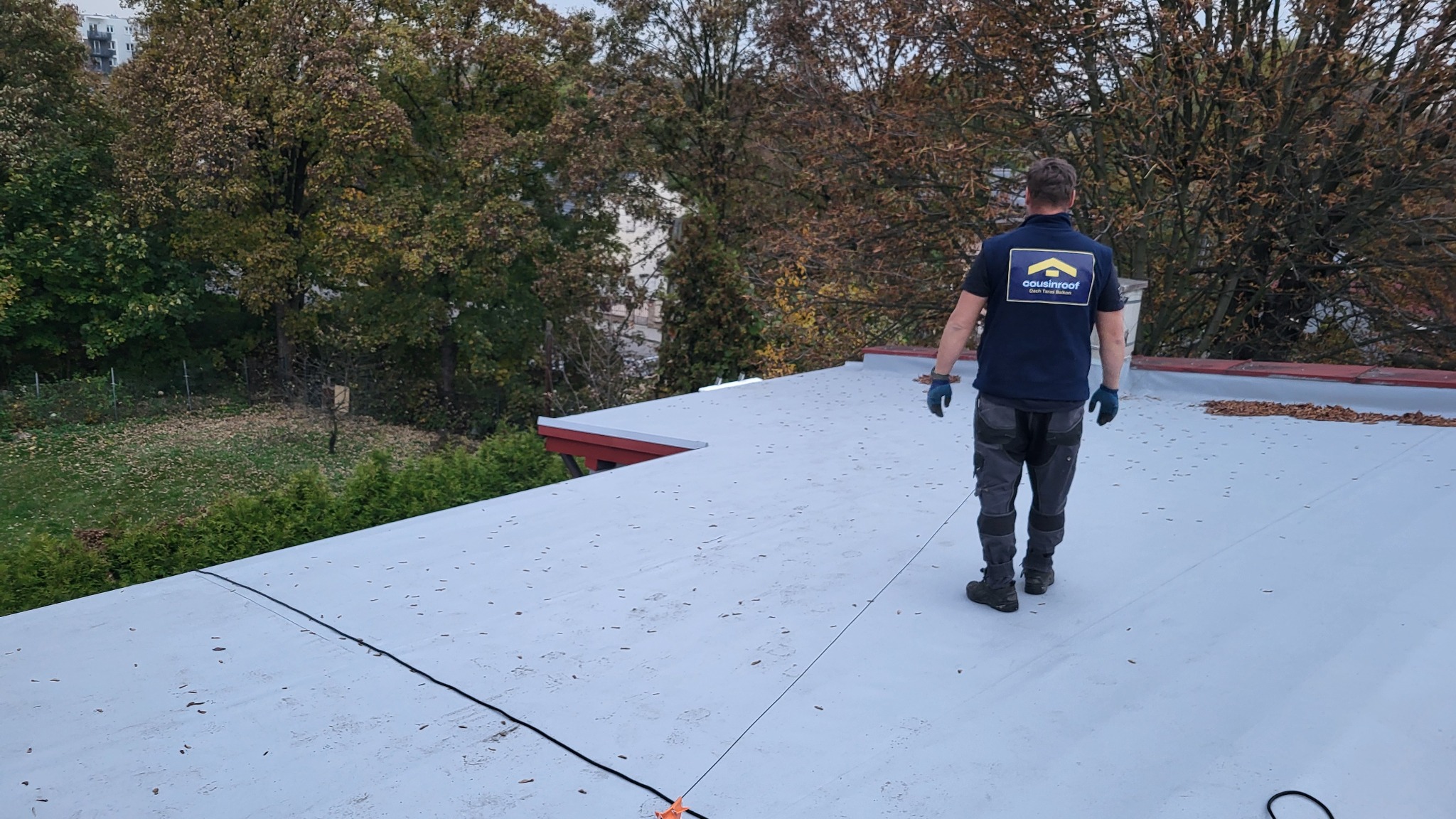 Pracownik w uniformie z logo firmy na płaskim dachu pokrytym membraną PVC, zabezpieczony linką asekuracyjną. W tle drzewa i budynki.