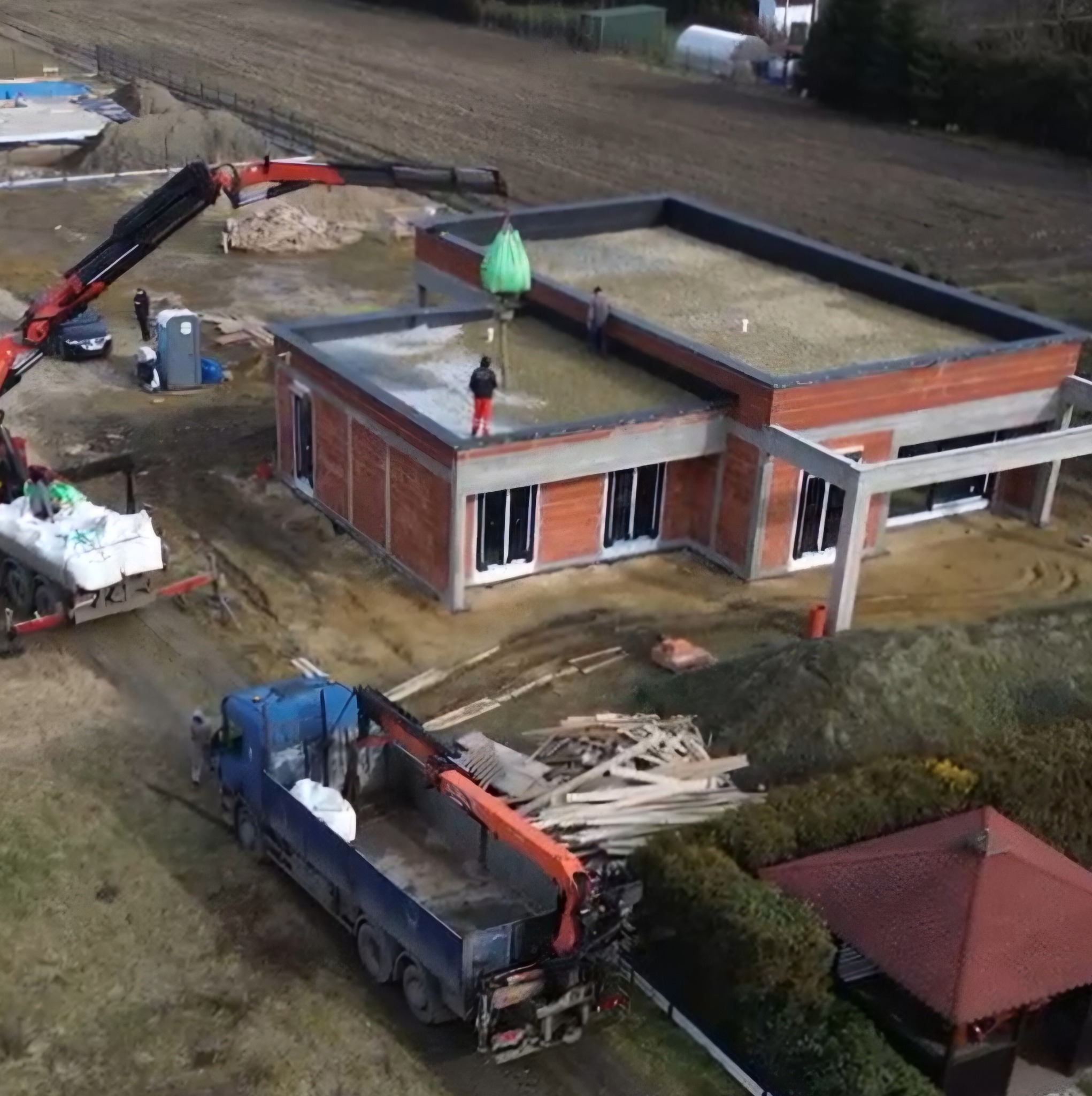 Budowa domu z płaskim dachem w trakcie prac: dźwig podnosi worki z materiałem na dach. Widok z góry na plac budowy i częściowo wykończony budynek.