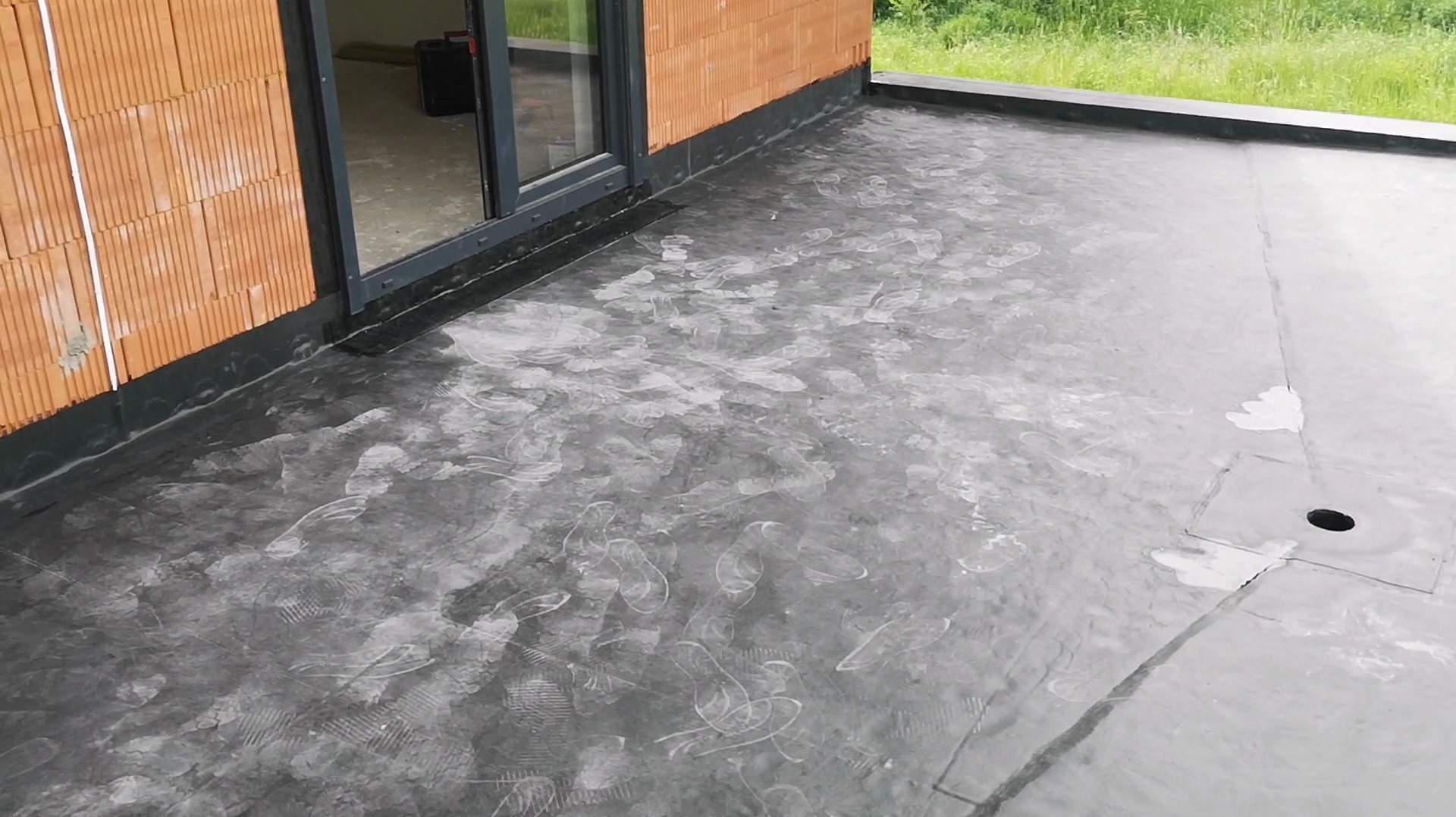 Wykończony dach płaski z odpływem, pokryty hydroizolacją z widocznymi śladami stóp, przylegający do ściany budynku z pustaków ceramicznych i okna.