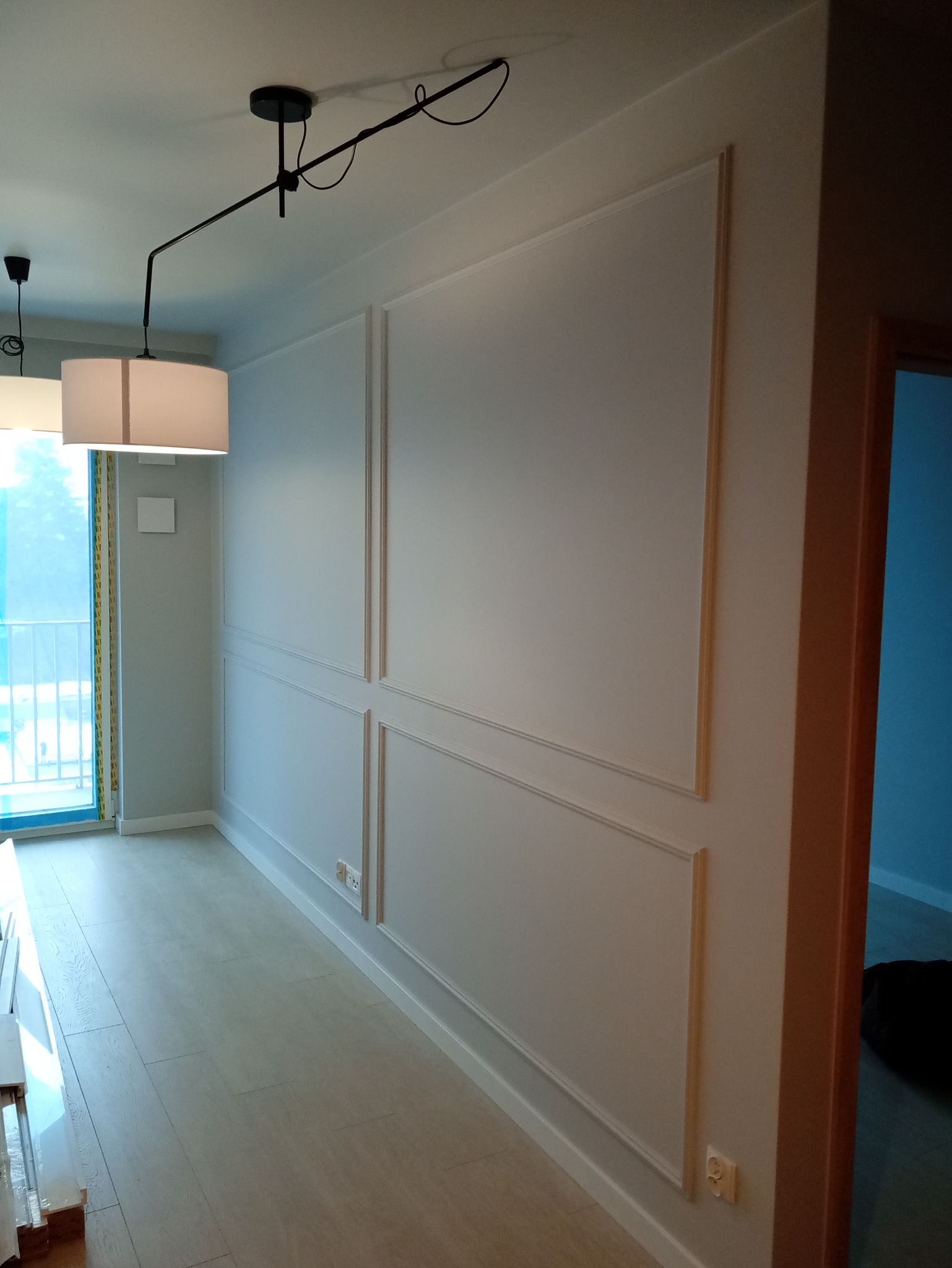 Białe ściany z dekoracyjnymi listwami w nowym mieszkaniu, widok na podłogę i okno balkonowe. Minimalistyczna lampa sufitowa. Gniazdka elektryczne na ścianie.