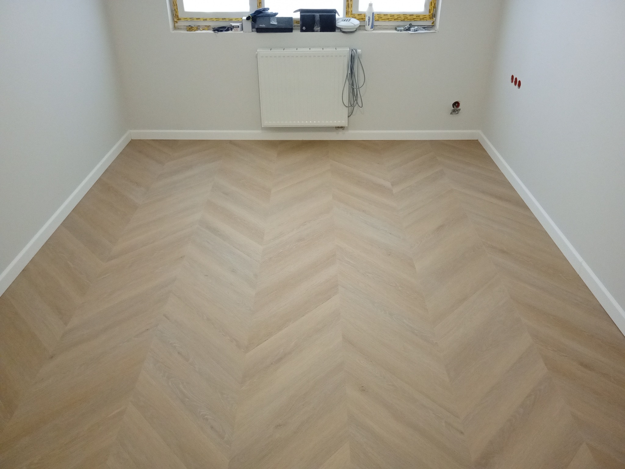 Jasna podłoga ułożona w jodełkę w pustym pokoju z grzejnikiem pod oknem. Widoczne listwy przypodłogowe i gniazdka elektryczne.