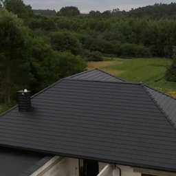 Mariusz Wawszczyk WAW-BUD - Czarny, nowy dach z kominem na tle zieleni. Widok z góry ukazuje regularne ułożenie dachówek i estetyczne wykończenie. Nowoczesny design wkomponowany w naturalny krajobraz.