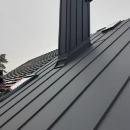 Mariusz Wawszczyk WAW-BUD - Grafitowy dach z blachy na rąbek stojący z kominem i oknami dachowymi, widok z dołu, fragment dachu w trakcie prac dekarskich z widoczną izolacją.
