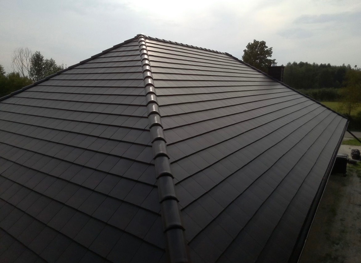 Ciemnoszary dach z dachówki ceramicznej, widok z góry, z wyraźnie zaznaczonym kalenicą i rynną. W tle zieleń drzew i pochmurne niebo.