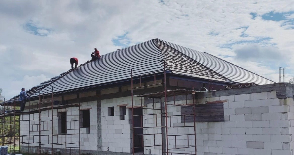 Układanie ciemnej dachówki na skośnym dachu domu jednorodzinnego. Prace dekarskie w toku, widoczne rusztowania i pracownicy na dachu. Budynek w stanie surowym.