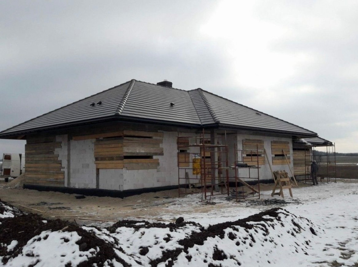 Dom w trakcie budowy z szarą dachówką, zabezpieczonymi oknami i rusztowaniami. W tle zimowy krajobraz i pochmurne niebo. Widoczny fragment elewacji.