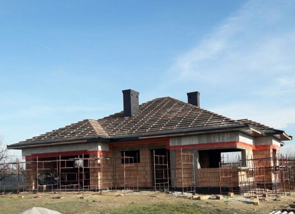 Niedokończony dom jednorodzinny z ciemną dachówką ceramiczną i dwoma kominami. Widoczne rusztowania i niezabudowane okna. Błękitne niebo w tle.