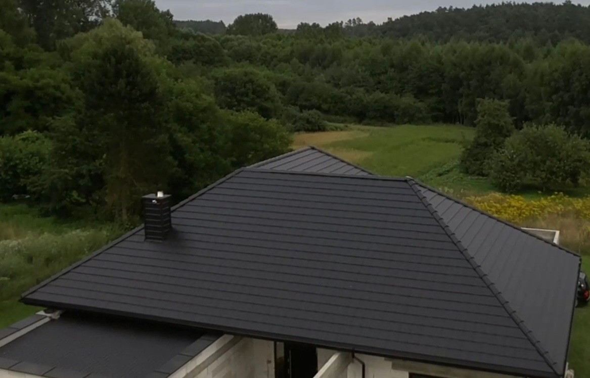 Czarny, nowy dach z kominem na tle zieleni. Widok z góry ukazuje regularne ułożenie dachówek i estetyczne wykończenie. Nowoczesny design wkomponowany w naturalny krajobraz.