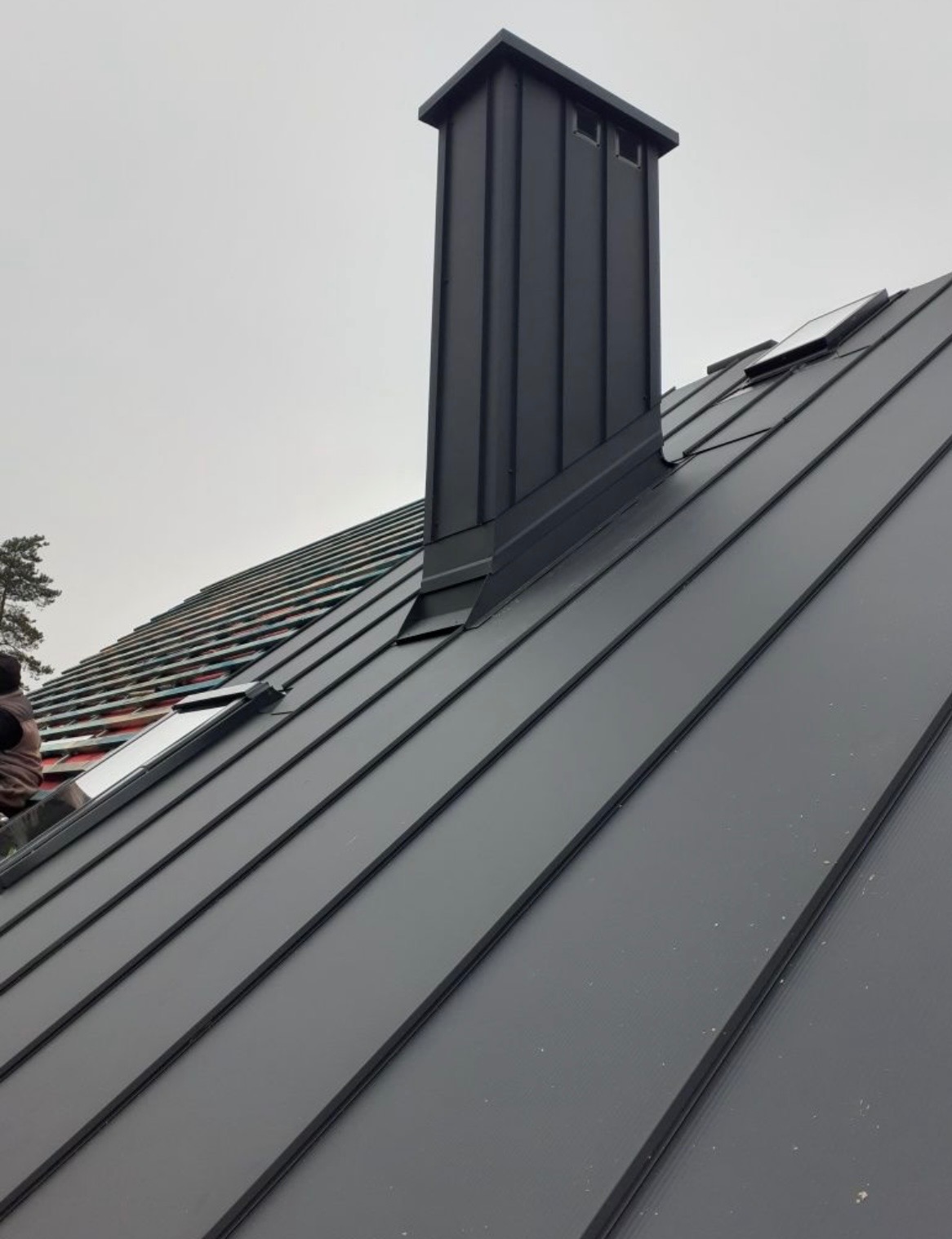 Grafitowy dach z blachy na rąbek stojący z kominem i oknami dachowymi, widok z dołu, fragment dachu w trakcie prac dekarskich z widoczną izolacją.