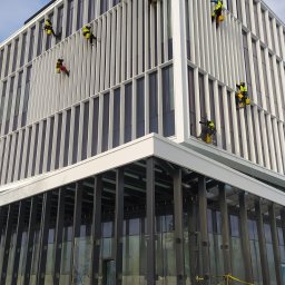 MMHigh - Ekipa alpinistów w jaskrawych strojach czyści szklaną fasadę nowoczesnego biurowca w zimowej scenerii, widok z dołu na tle błękitnego nieba.