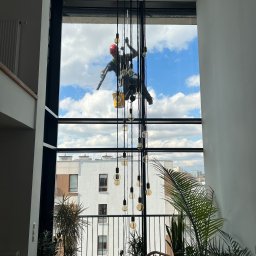 MMHigh - Pracownik w kasku, na linach, myje okno w wysokim apartamencie. Widok na błękitne niebo i budynki. Wnętrze z roślinami i lampami wiszącymi.