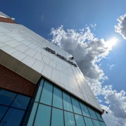 MMHigh - Pracownik na wysokości myje elewację biurowca z logo, widoczne liny i sprzęt alpinistyczny. Błękitne niebo z chmurami w tle. Ujęcie z dołu.