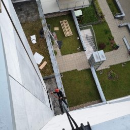 MMHigh - Pracownik w kasku, na linach, wykonuje prace wysokościowe na elewacji budynku. Widok z góry, z perspektywy liny. W tle dziedziniec z zielenią i elementami architektury.