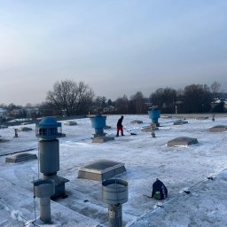 MMHigh - Osoba odśnieża płaski dach pokryty śniegiem, widoczne kominy wentylacyjne i świetliki dachowe. W tle zabudowa miejska i drzewa.