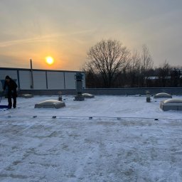 MMHigh - Pracownik odśnieża płaski dach budynku komercyjnego przy zachodzącym słońcu. Widoczne świetliki dachowe i drzewa w tle. Zima.