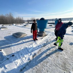 MMHigh - Ekipa w zimowych ubraniach na zaśnieżonym dachu z świetlikami i wentylacją. Ślady odśnieżania widoczne na powierzchni. Błękitne niebo w tle.
