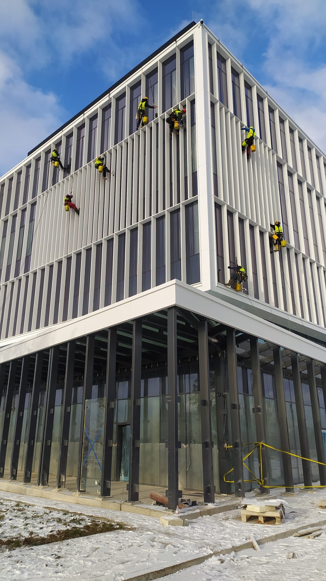 Ekipa alpinistów w jaskrawych strojach czyści szklaną fasadę nowoczesnego biurowca w zimowej scenerii, widok z dołu na tle błękitnego nieba.