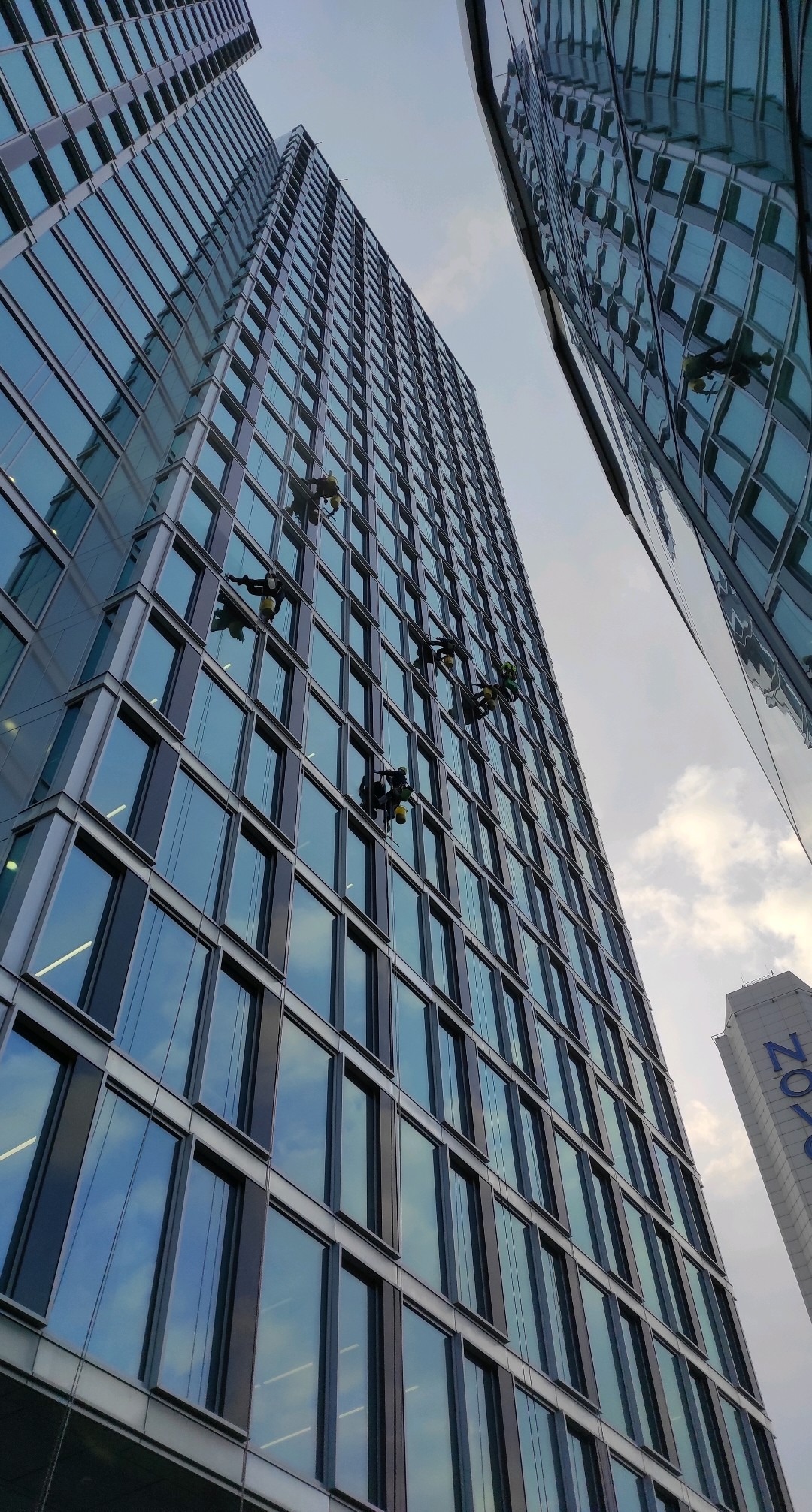 Wysokiej klasy budynek biurowy z pracownikami alpinistycznymi myjącymi okna na wysokości. Widok z dołu, pochmurne niebo w tle.