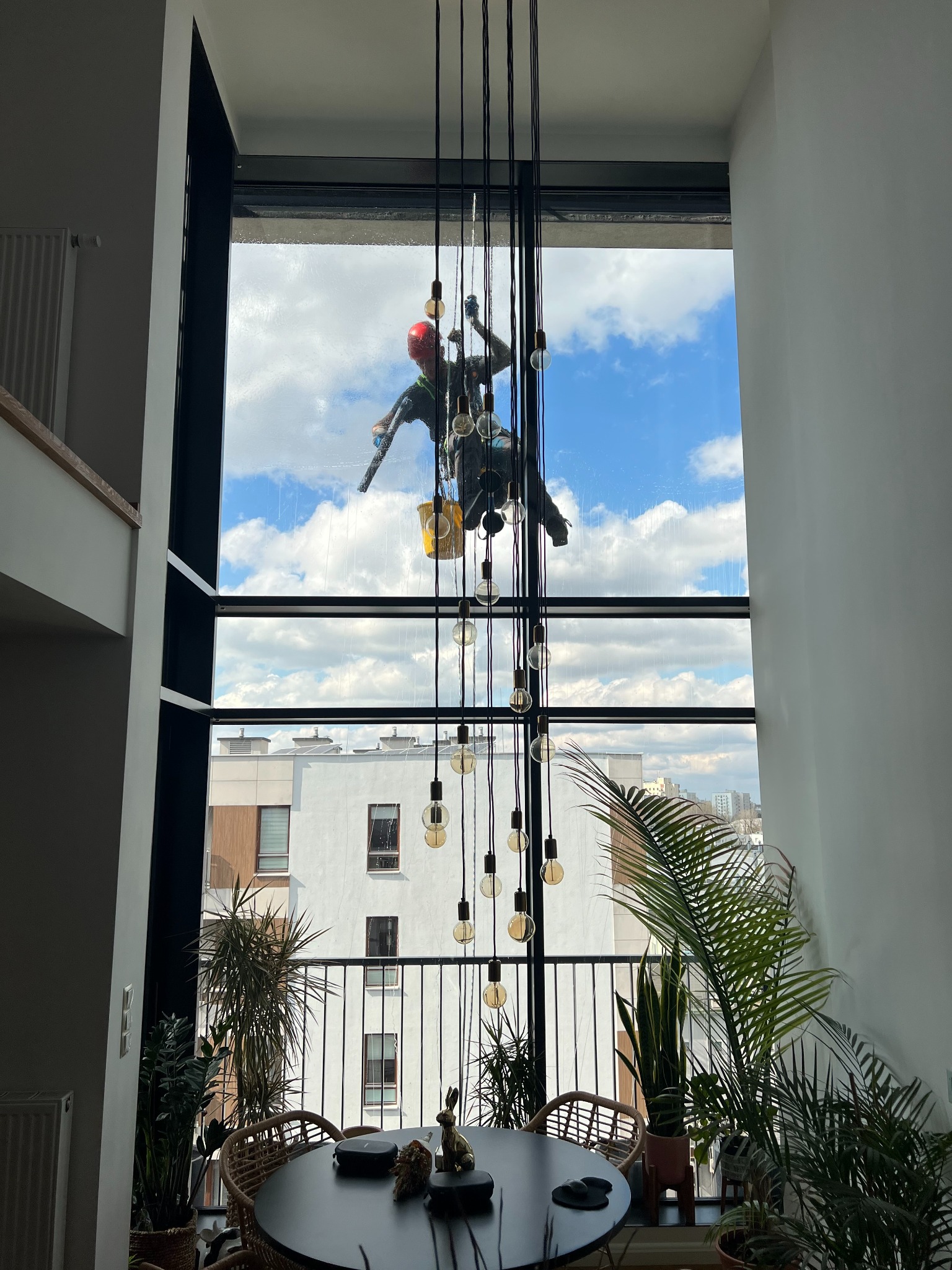 Pracownik w kasku, na linach, myje okno w wysokim apartamencie. Widok na błękitne niebo i budynki. Wnętrze z roślinami i lampami wiszącymi.