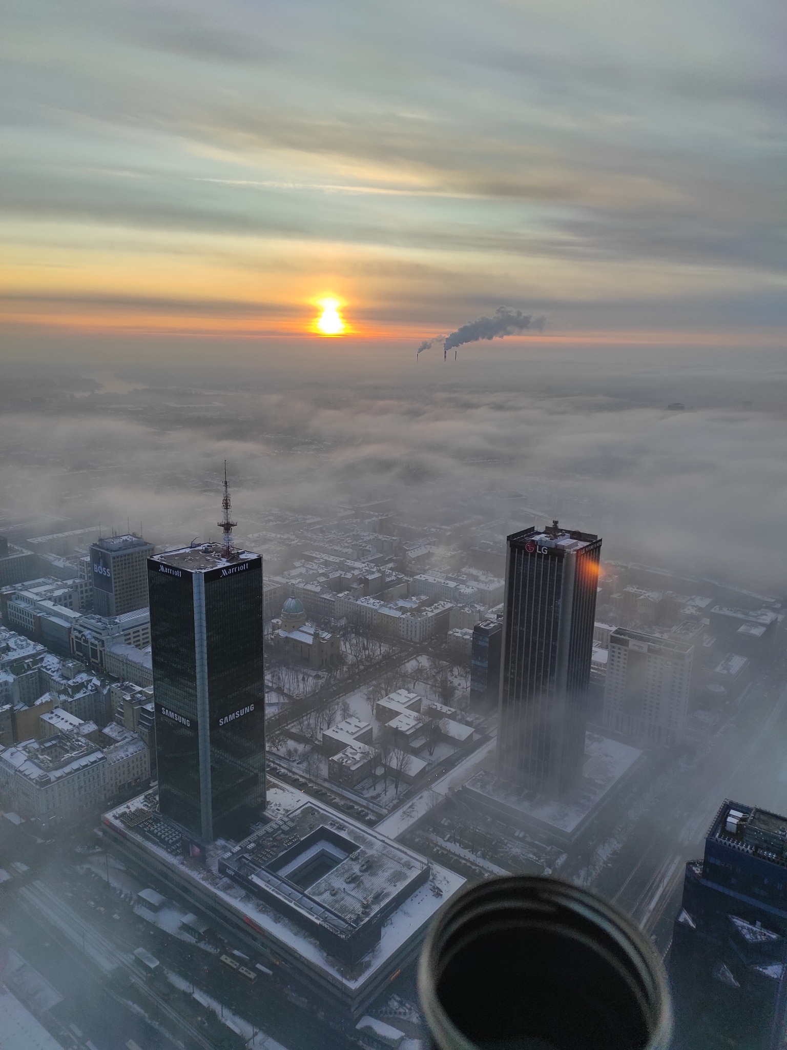 Zimowy widok z wysokości na Warszawę spowitą mgłą, z wieżowcami Samsung i LG, kominami w oddali i wschodzącym słońcem. Na pierwszym planie fragment metalowej konstrukcji.