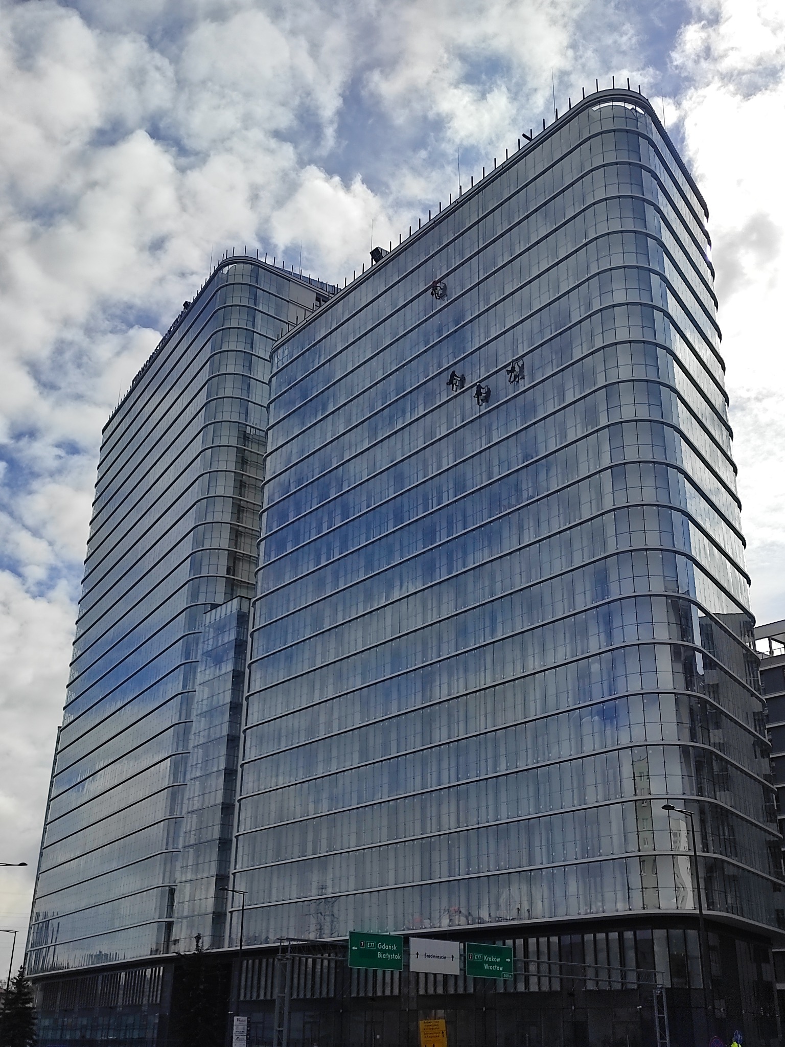 Dwaj pracownicy na wysokościach myją okna nowoczesnego, szklanego biurowca w słoneczny dzień. Widoczne odbicie chmur w fasadzie.