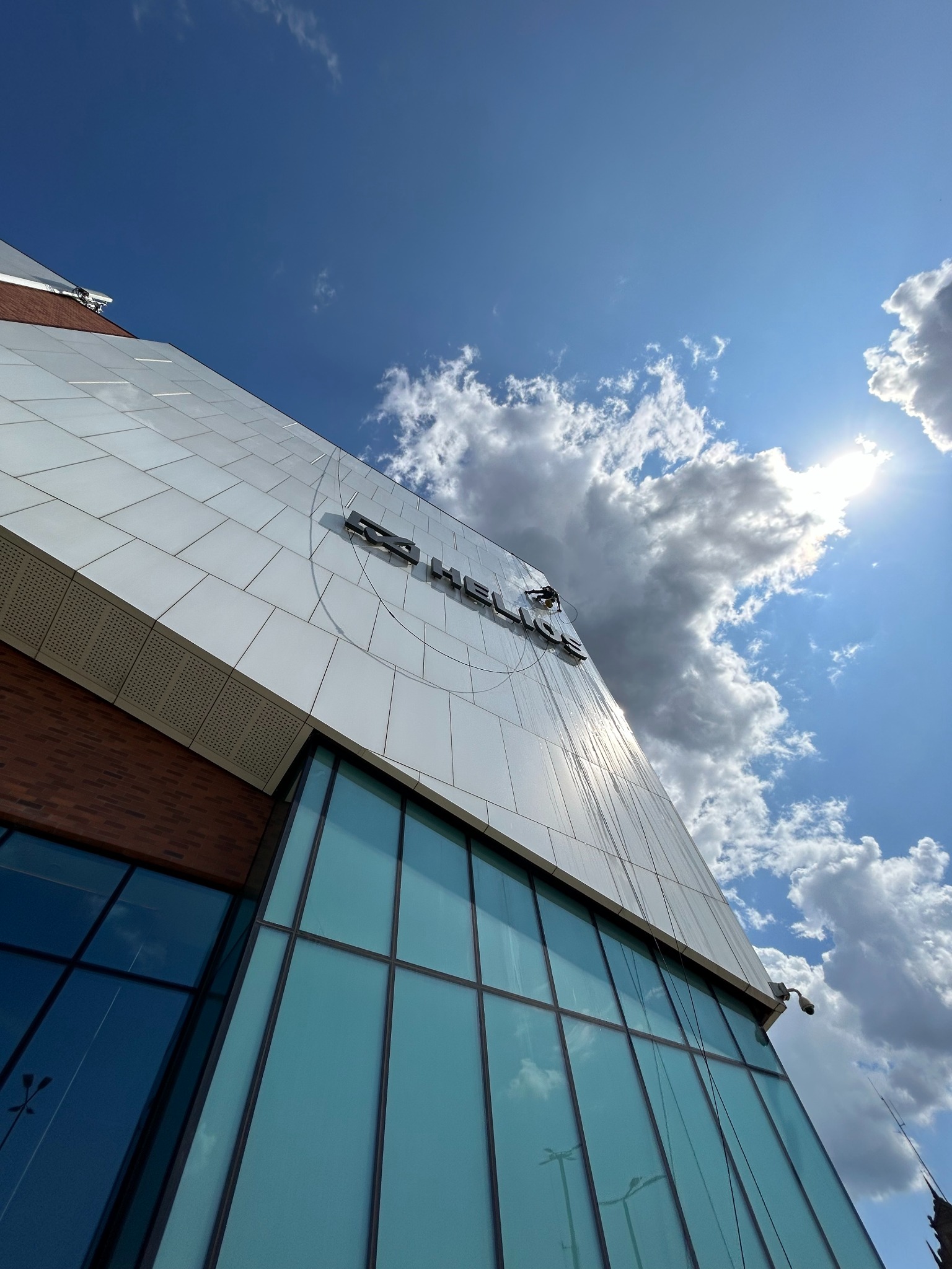 Pracownik na wysokości myje elewację biurowca z logo, widoczne liny i sprzęt alpinistyczny. Błękitne niebo z chmurami w tle. Ujęcie z dołu.