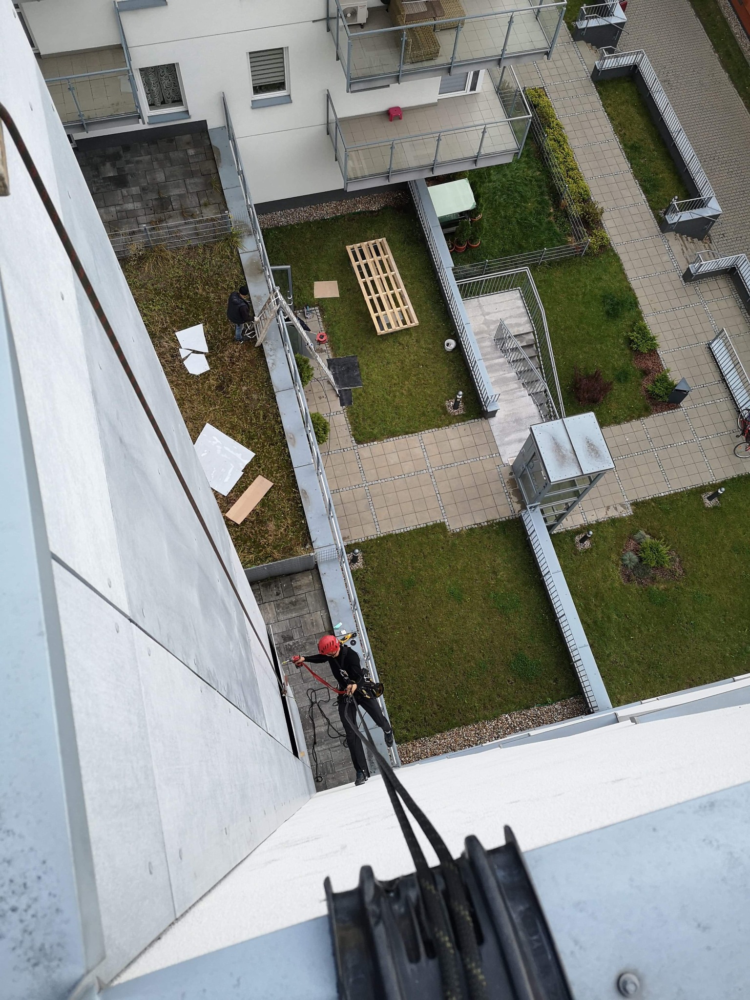 Pracownik w kasku, na linach, wykonuje prace wysokościowe na elewacji budynku. Widok z góry, z perspektywy liny. W tle dziedziniec z zielenią i elementami architektury.