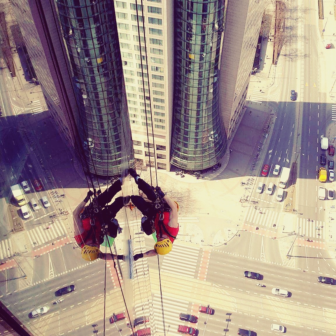 Dwóch alpinistów w kaskach czyści szklaną elewację wieżowca, widok z góry na miasto i ruch uliczny. Używają sprzętu do dostępu linowego.