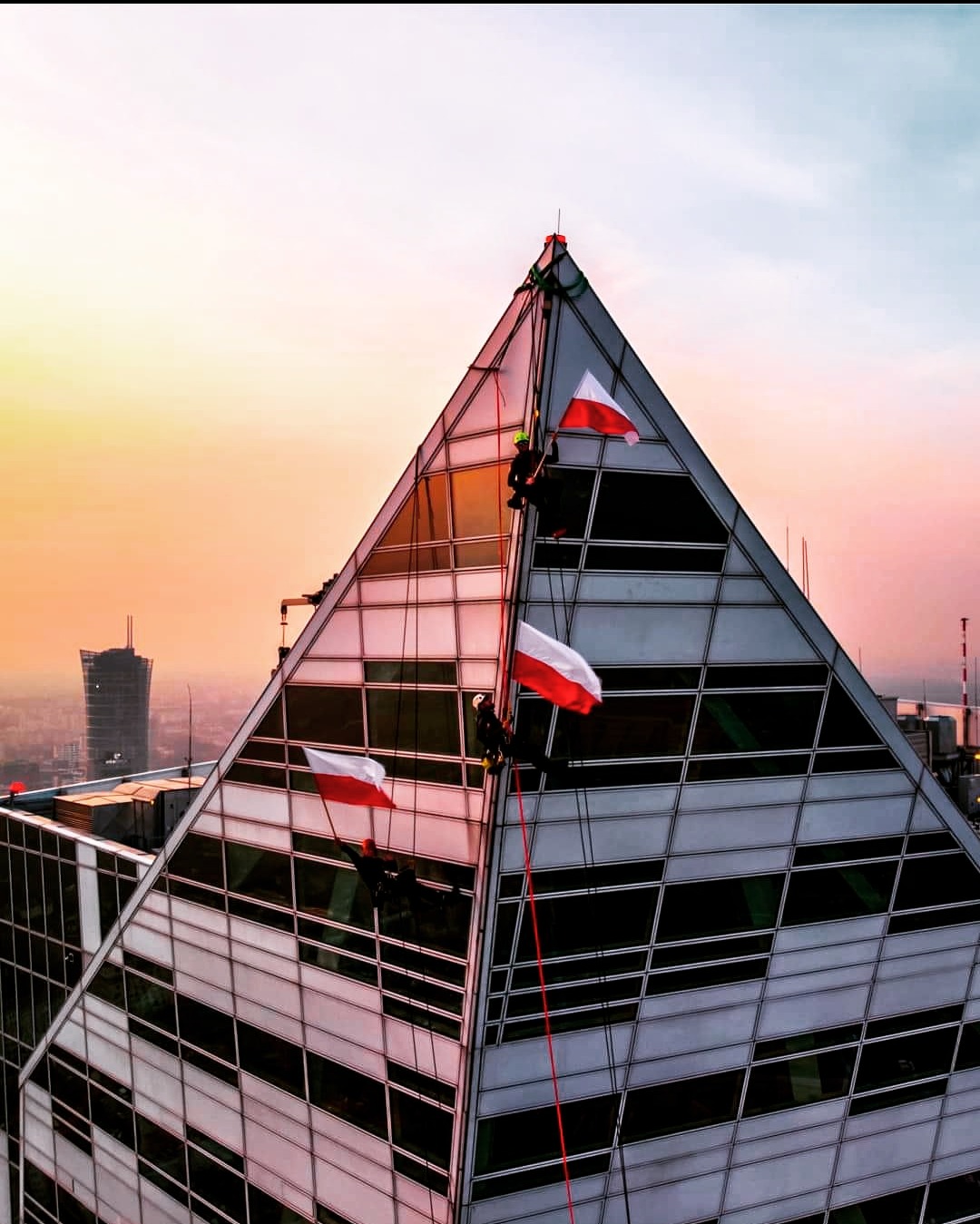 Alpiniści na szklanym budynku w kształcie piramidy, z flagami Polski na tle miejskiego krajobrazu o zachodzie słońca. Widok z dołu, liny i uprzęże.