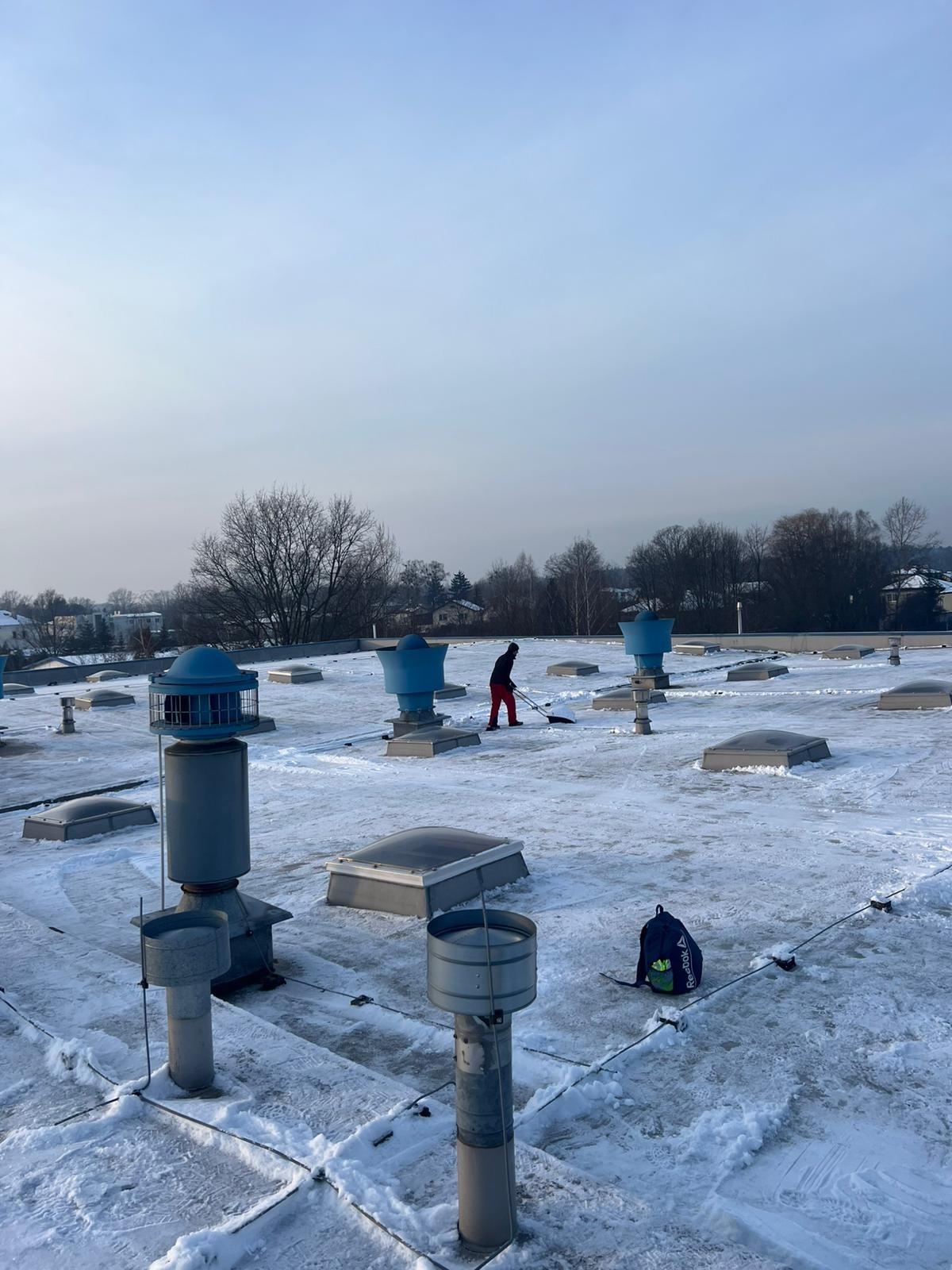 Osoba odśnieża płaski dach pokryty śniegiem, widoczne kominy wentylacyjne i świetliki dachowe. W tle zabudowa miejska i drzewa.