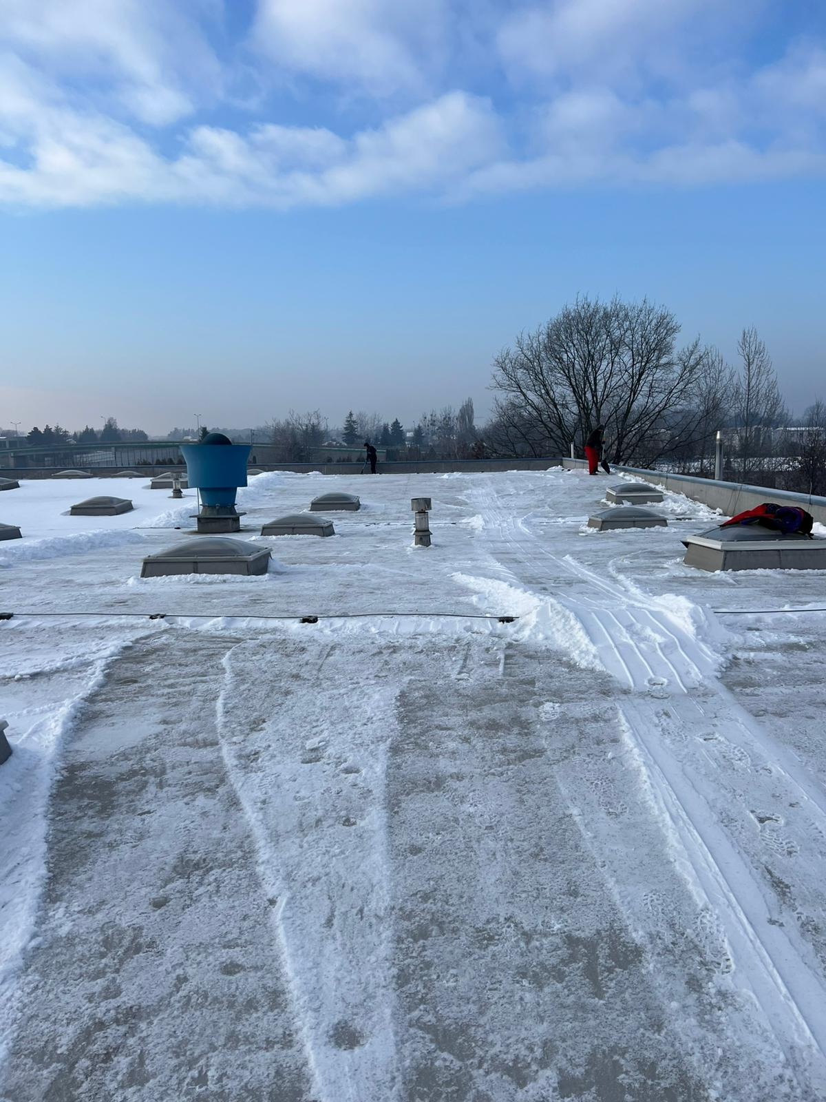 Odśnieżony dach płaski z widocznymi śladami odśnieżania, w tle zabudowa miejska i drzewa. Widoczne świetliki dachowe i elementy instalacji.