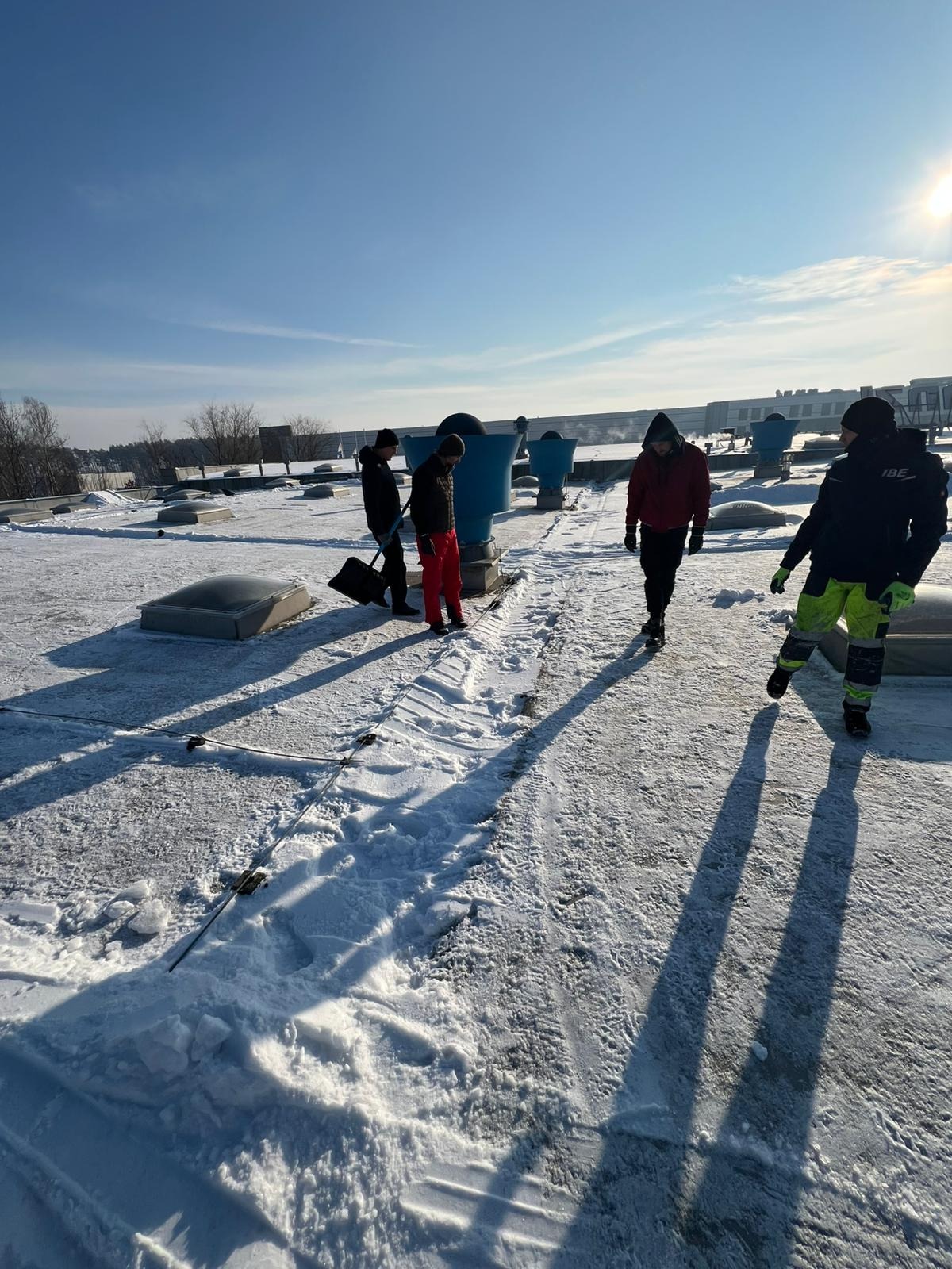 Ekipa w zimowych ubraniach na zaśnieżonym dachu z niebieskimi wentylatorami. Ślady odśnieżania widoczne, słoneczny dzień.