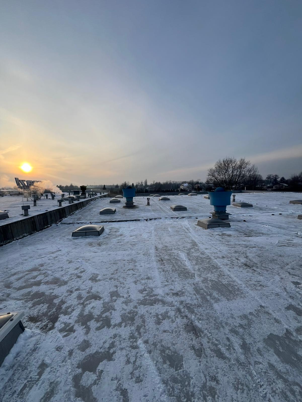 Śnieg na płaskim dachu z odpowietrznikami i świetlikami o poranku. Widoczne ślady częściowego odśnieżania, w tle zabudowania i drzewa.
