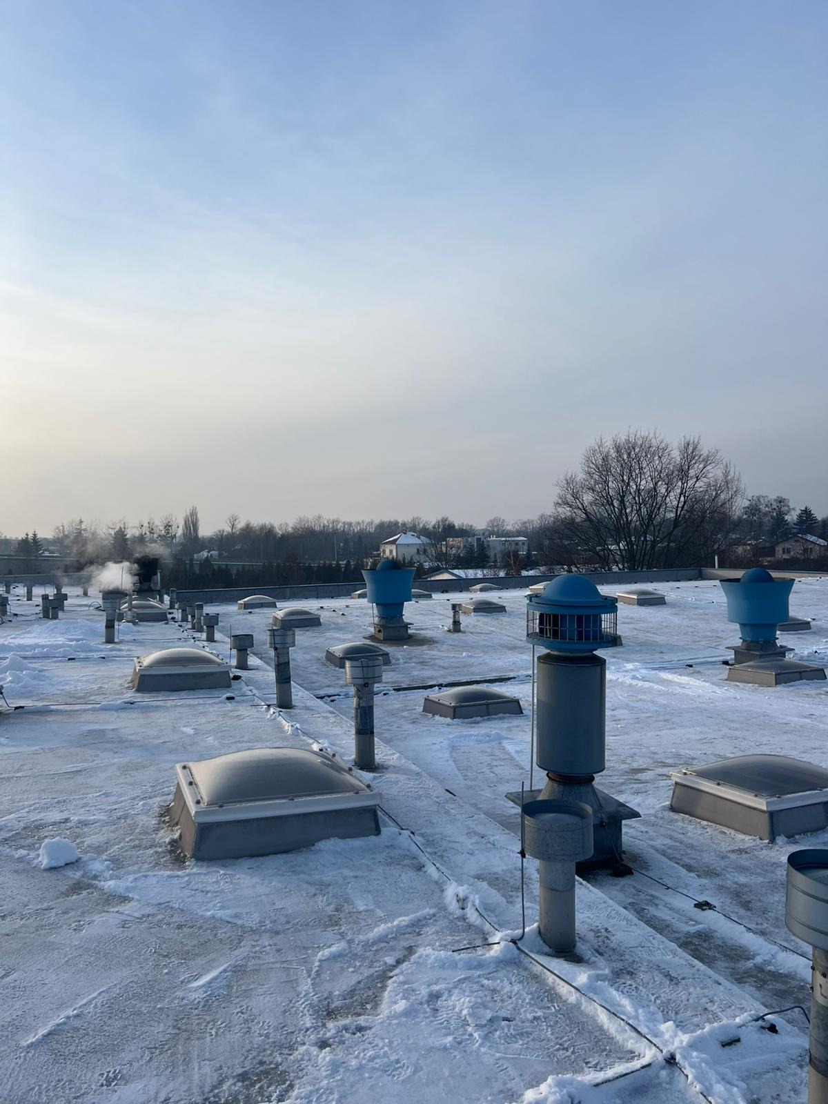 Śnieżny dach płaski z licznymi wywietrznikami i świetlikami, widok z góry w pochmurny dzień. Częściowo odśnieżony, z widocznymi śladami po odśnieżaniu.