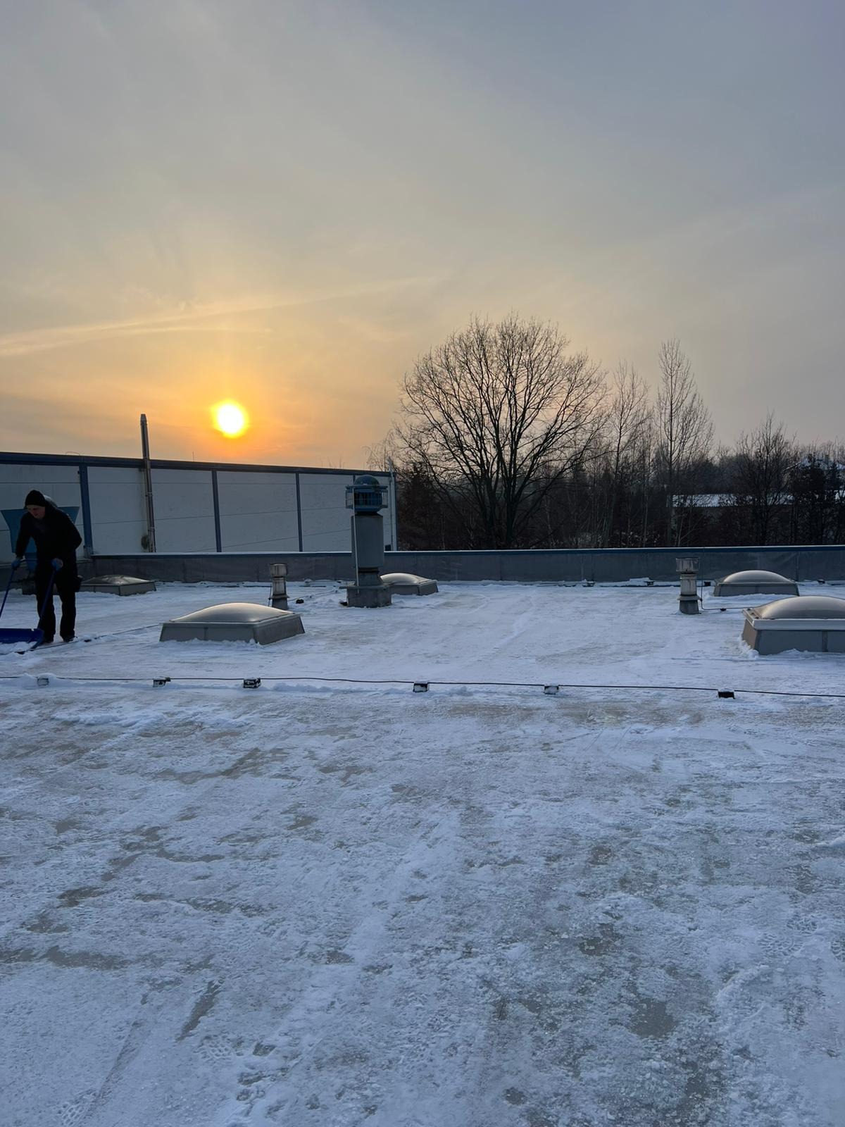 Pracownik odśnieża płaski dach budynku komercyjnego przy zachodzącym słońcu. Widoczne świetliki dachowe i drzewa w tle. Zima.