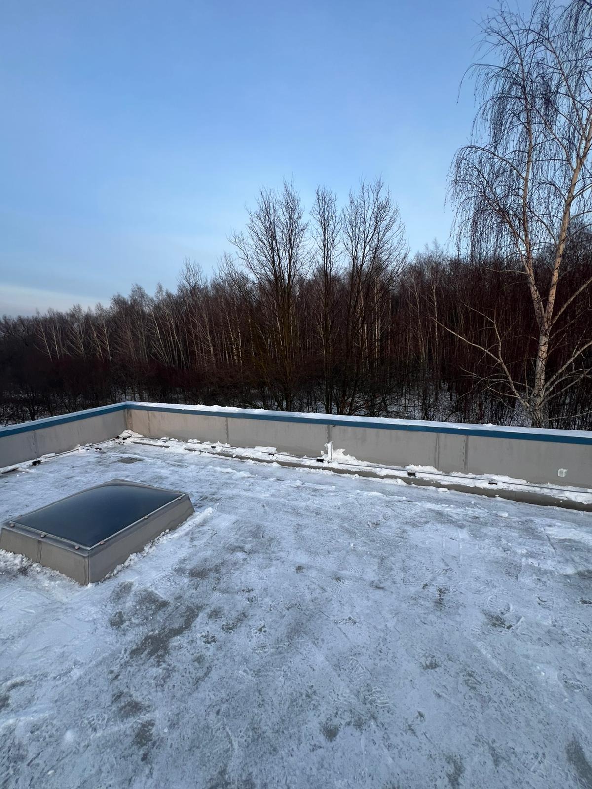 Zaśnieżony, płaski dach z świetlikiem w Kowalach, widok z góry. W tle drzewa bez liści i błękitne niebo. Śnieg częściowo stopniał, odsłaniając powierzchnię dachu.