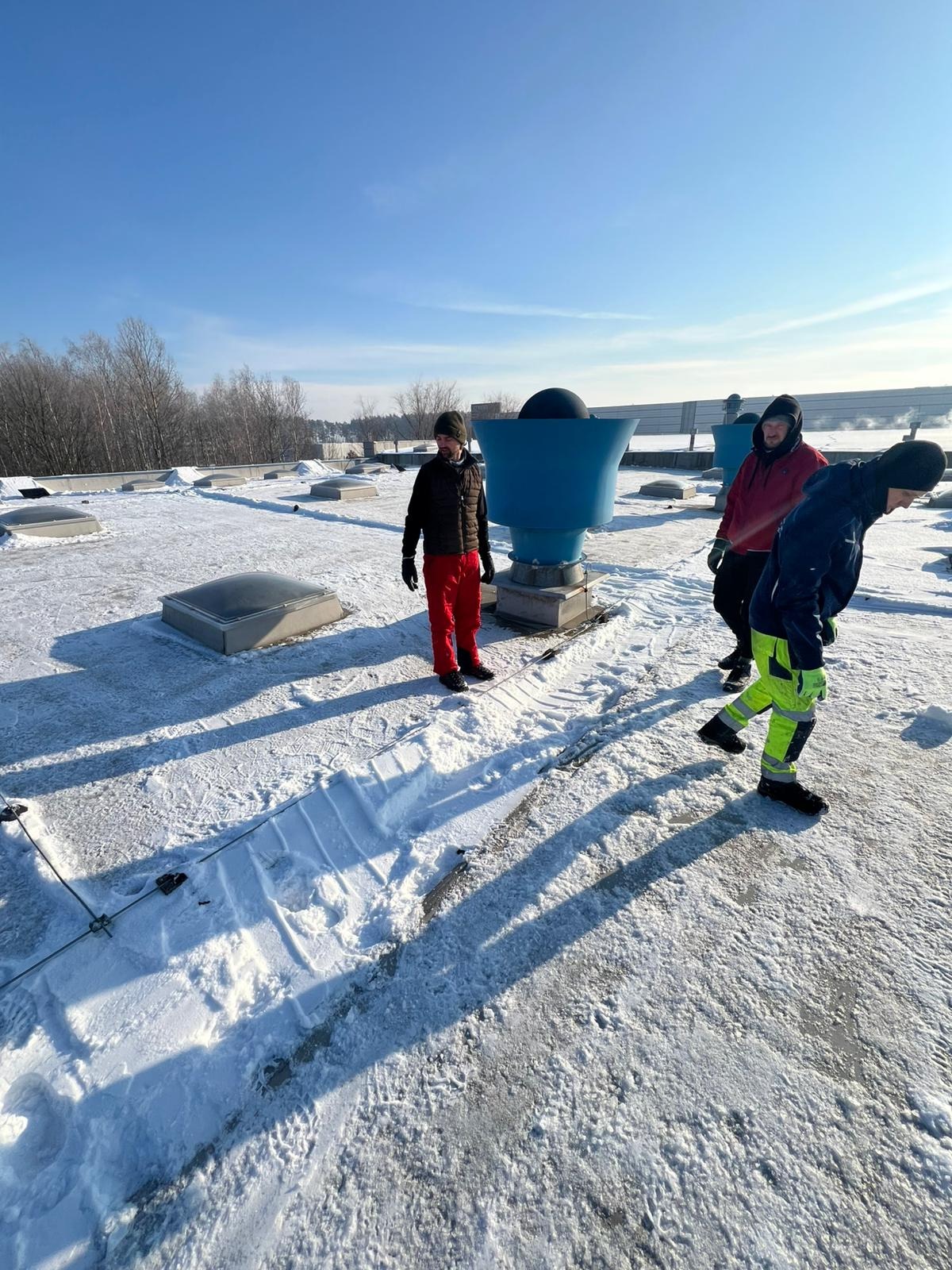 Ekipa w zimowych ubraniach na zaśnieżonym dachu z świetlikami i wentylacją. Ślady odśnieżania widoczne na powierzchni. Błękitne niebo w tle.