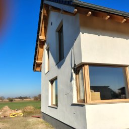 Tomasz Adamski - Elewacja domu jednorodzinnego w trakcie prac wykończeniowych, widoczne okna z drewnianą stolarką i fragment elewacji w szarym kolorze. W tle zielone pole i błękitne niebo.