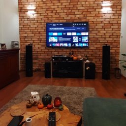 Tomasz Adamski - Ściana z cegły klinkierowej za telewizorem, oświetlona kinkietami. Element wykończenia wnętrza salonu z drewnianą podłogą i stolikiem kawowym.