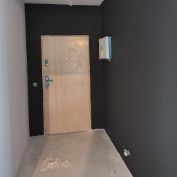 Tomasz Adamski - Świeżo pomalowane na czarno ściany w pokoju z surową podłogą i zabezpieczonymi elementami. Widoczna drewniana ościeżnica i drzwi. Etap prac wykończeniowych.